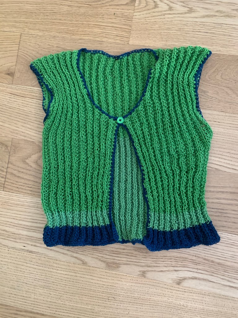 The front of a green knit vest with blue hem and crochet edging and a single green button at the center neck laying on a light wood floor.