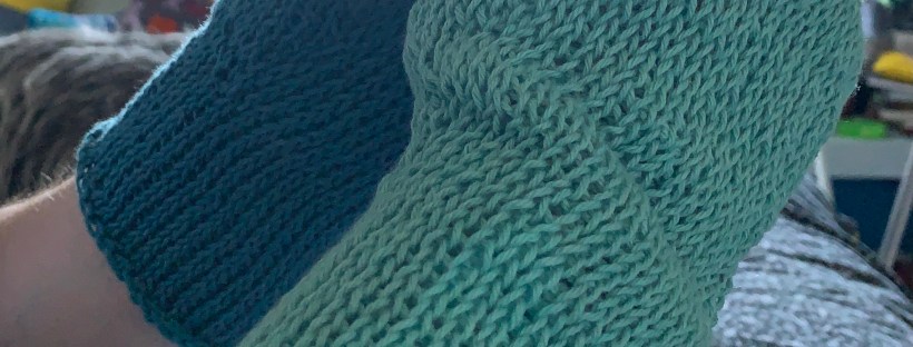 Two hands in blue Tunisian crocheted gloves holding a coffee cup, slightly backlit.