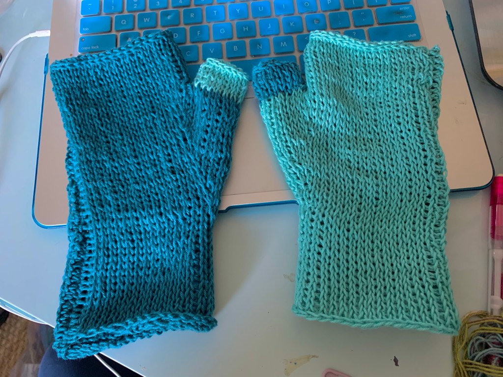 A pair of blue Tunisian crocheted gloves lay on a laptop keyboard. One is dark blue with a light blue thumb and the other is light blue with a dark blue thumb.