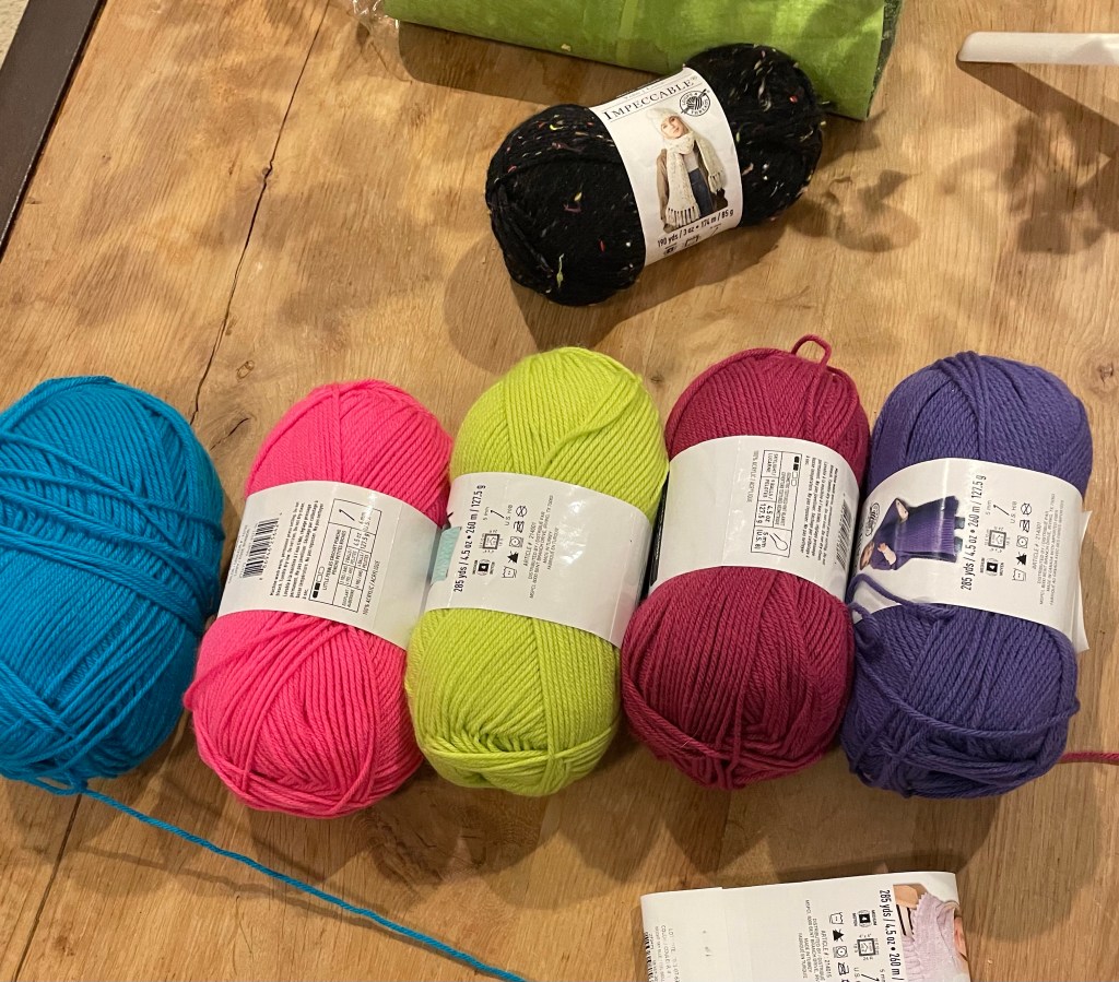 Five skeins of neon yarn on a light wooden table: blue, pink, green, magenta, and purple from left to right. A speckled black skein sit above them.