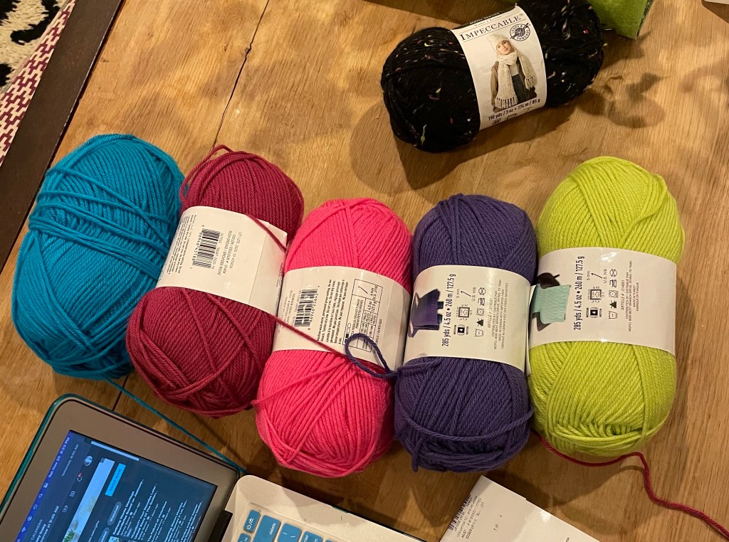 Five skeins of neon yarn on a light wooden table: blue, magenta, pink, purple, and magenta from left to right. A speckled black skein sit above them.