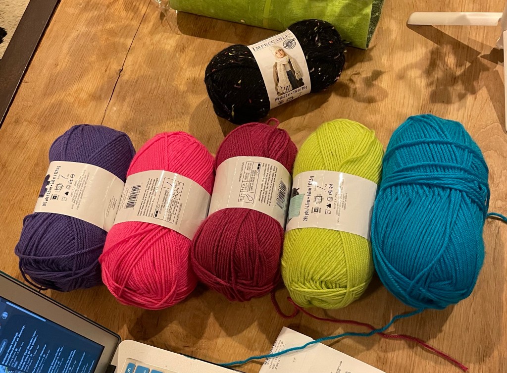 Five skeins of neon yarn on a light wooden table: purple, pink, magenta, green, and blue from left to right. A speckled black skein sit above them.