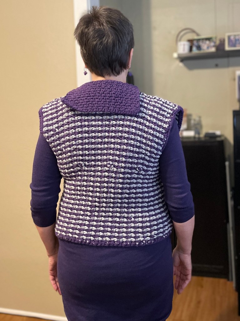 A white woman with short brown hair models a purple and gray striped crochet vest, facing away from the camera to show off the back and the purple collar.
