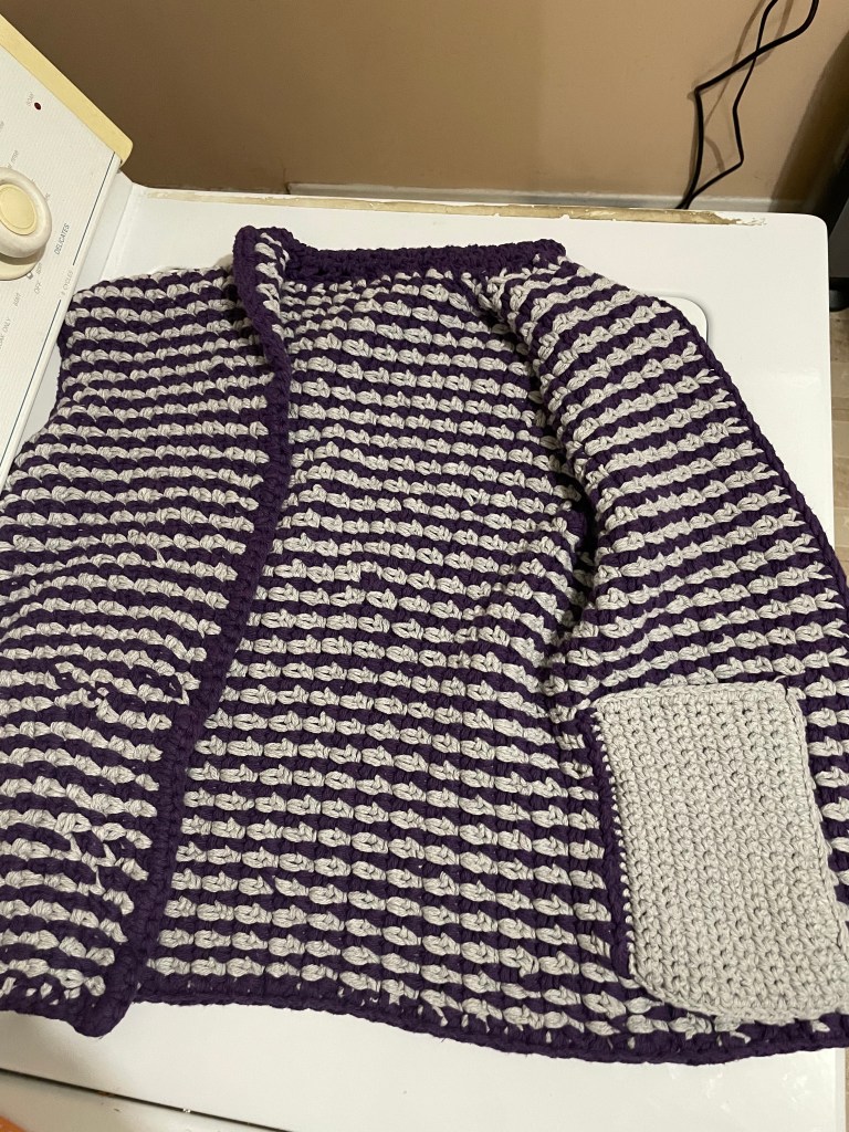 A purple and gray striped crochet vest laying flat wth one side open, showing off the gray pocket.