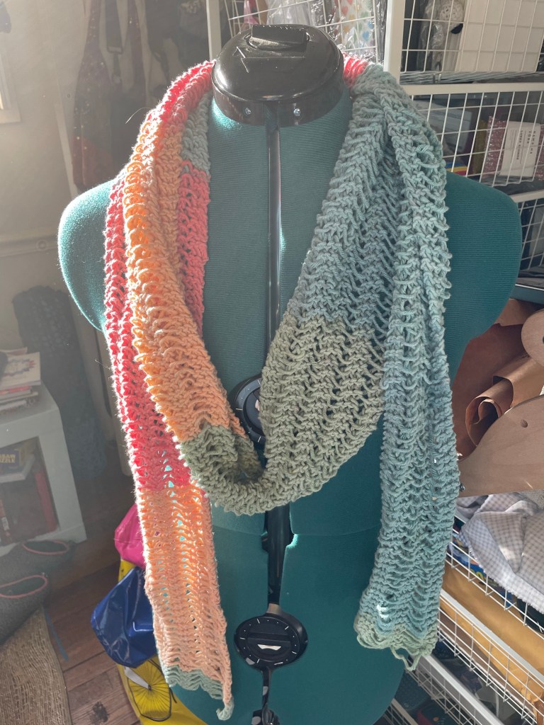 A green, blue, orange, and red knit scarf with dropped stitch details draped around the neck of a green, adjustable dress form.