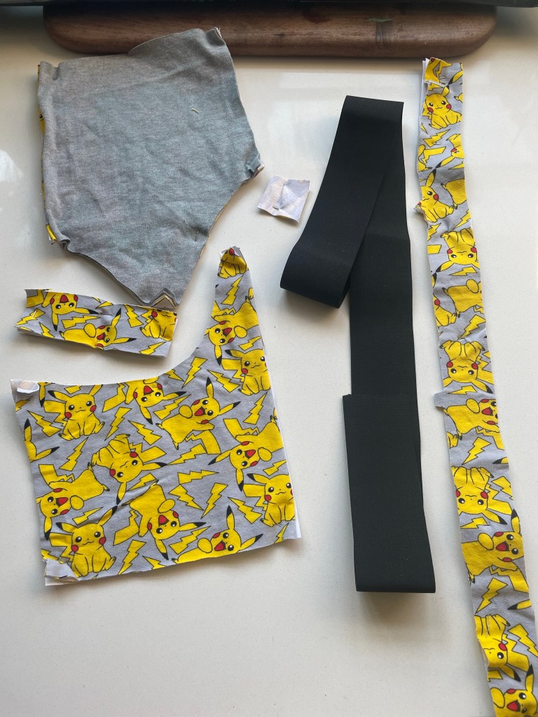 Pattern pieces of a bralette cut out of gray fabric with Pikachu printed all over and a strip of black elastic laid out on a white counter.