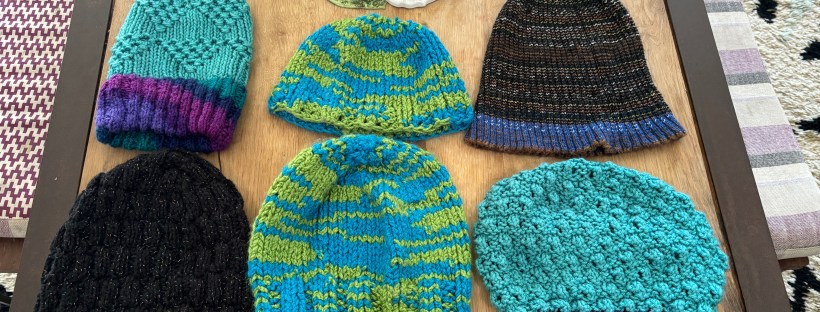 Six knit hats laid out in two rows of three. From top left: light blue body with triangles of textured stitches and a ribbed brim in shades of purple, a plain hat with puddles of green and blue, a brown and black striped ribbed beanie with blue stripes in the brim, a bubble-stitch teal hat with black and neon blue, purple, green, and pink striped brim, another green and blue hat from the same yarn as the one above it, and a sparkled black hat with short teal brim.
