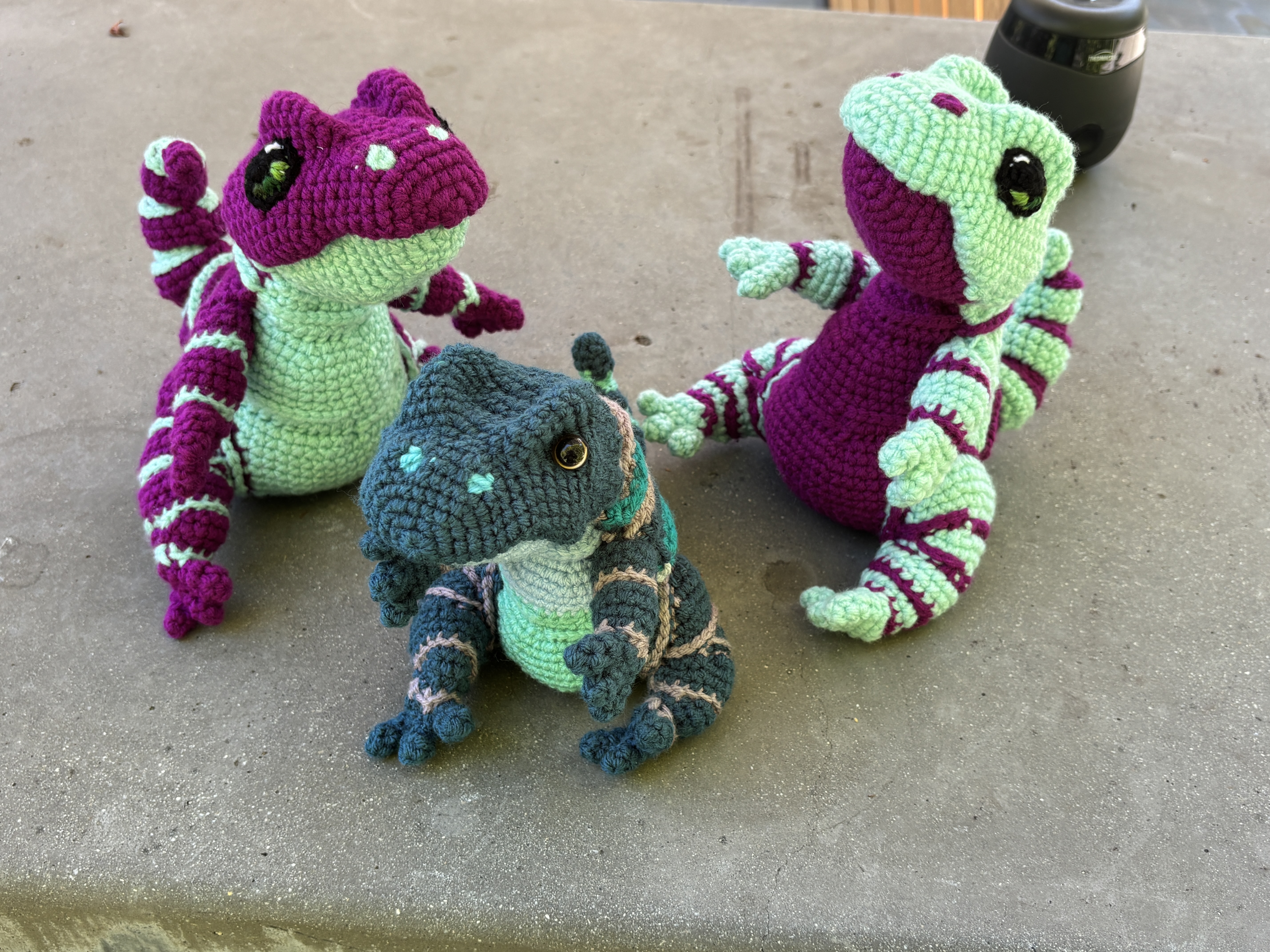 A group of three  crocheted baby dinosaurs. The top two have inverse coloring in light mint and dark maroon with crocheted eyes and the bottom onf has dark and light teal with gray accents and black safety eyes.