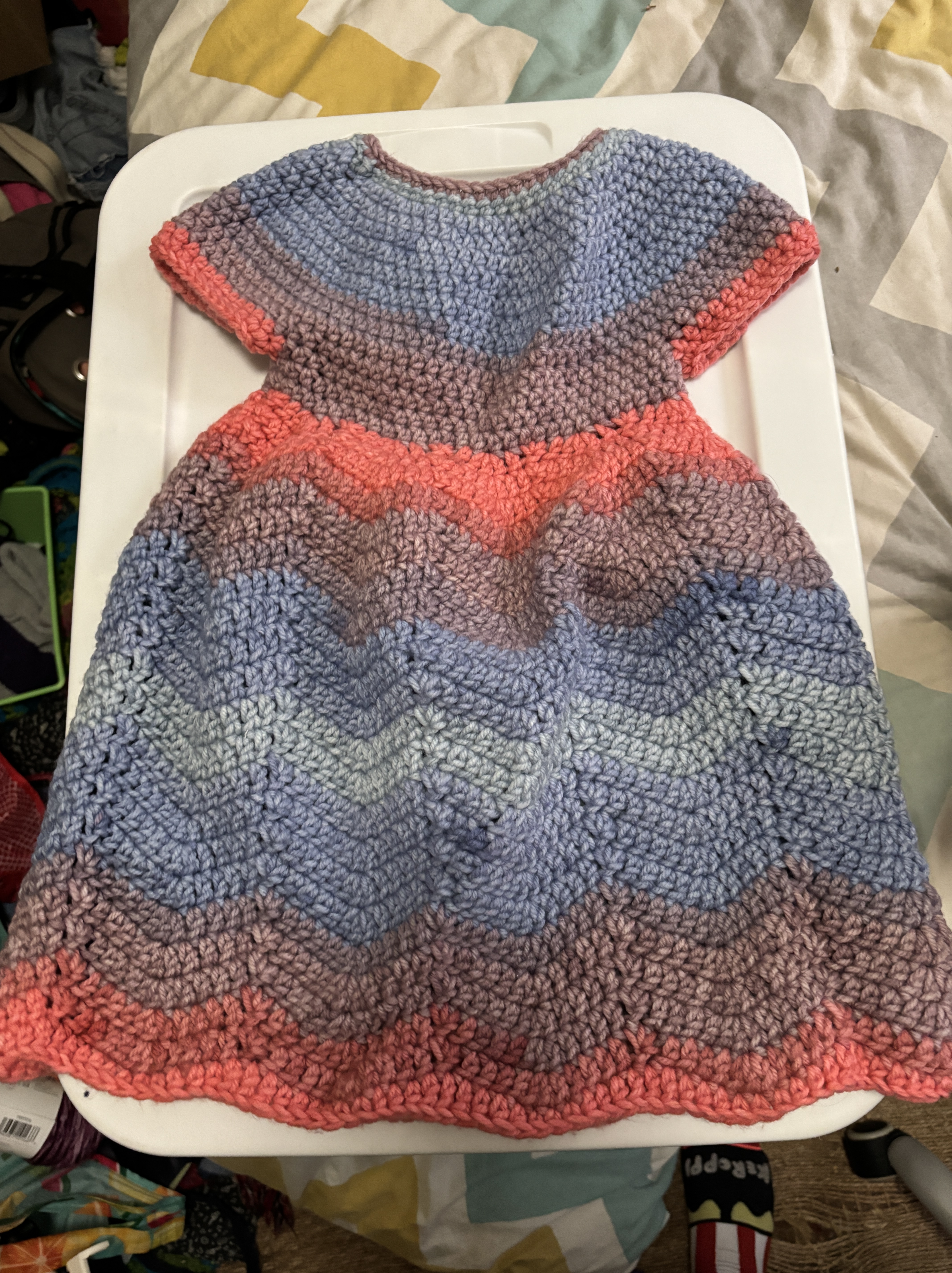 A child sized crochet dress with stripes of pink, blue, and purple and a chevron stitch pattern to the skirt.