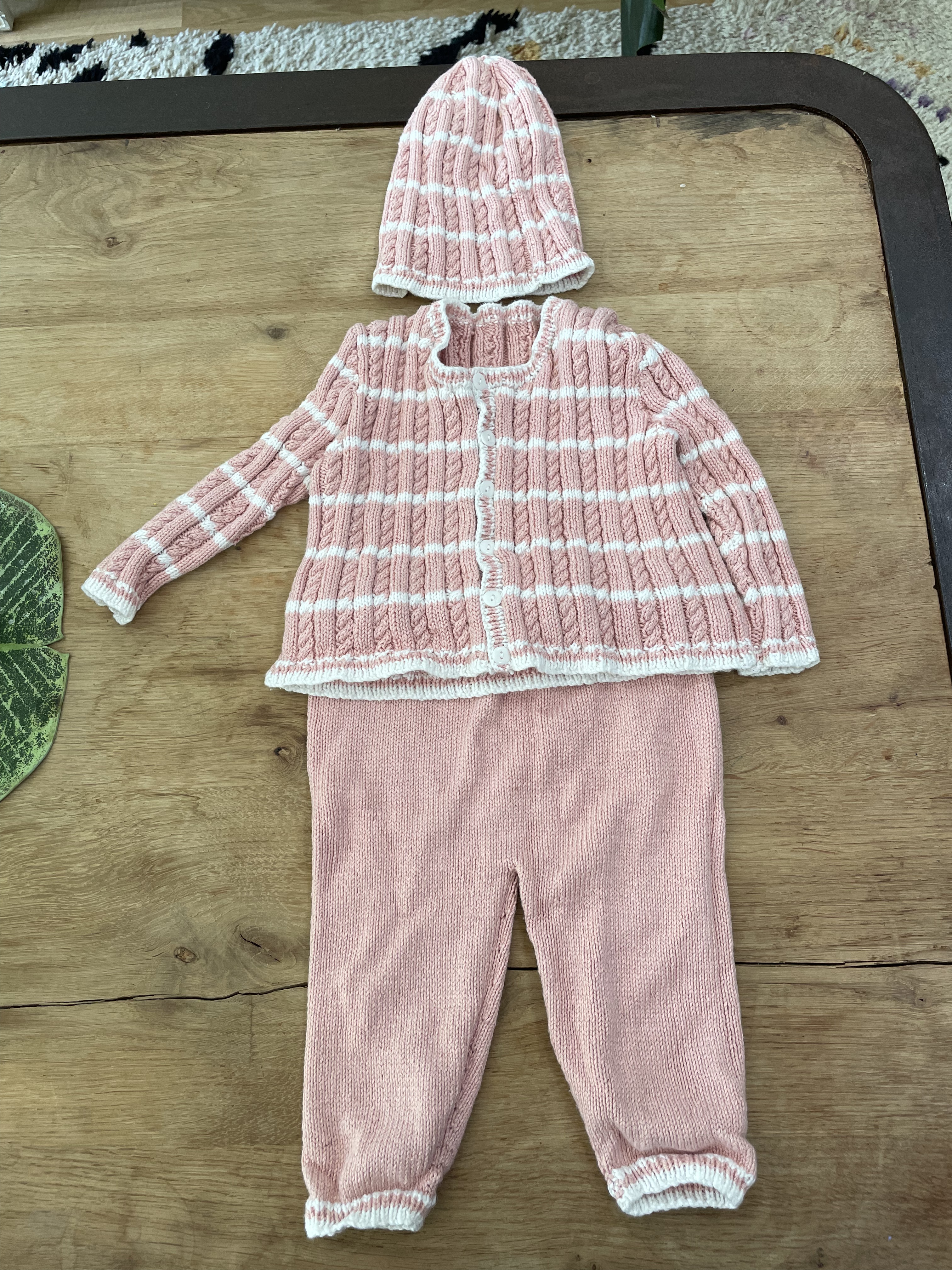 A pink and white striped baby set featuring a plain knit pair of pants, a cabled and textured jacket with white buttons, and a hat witht he same cables and patterning