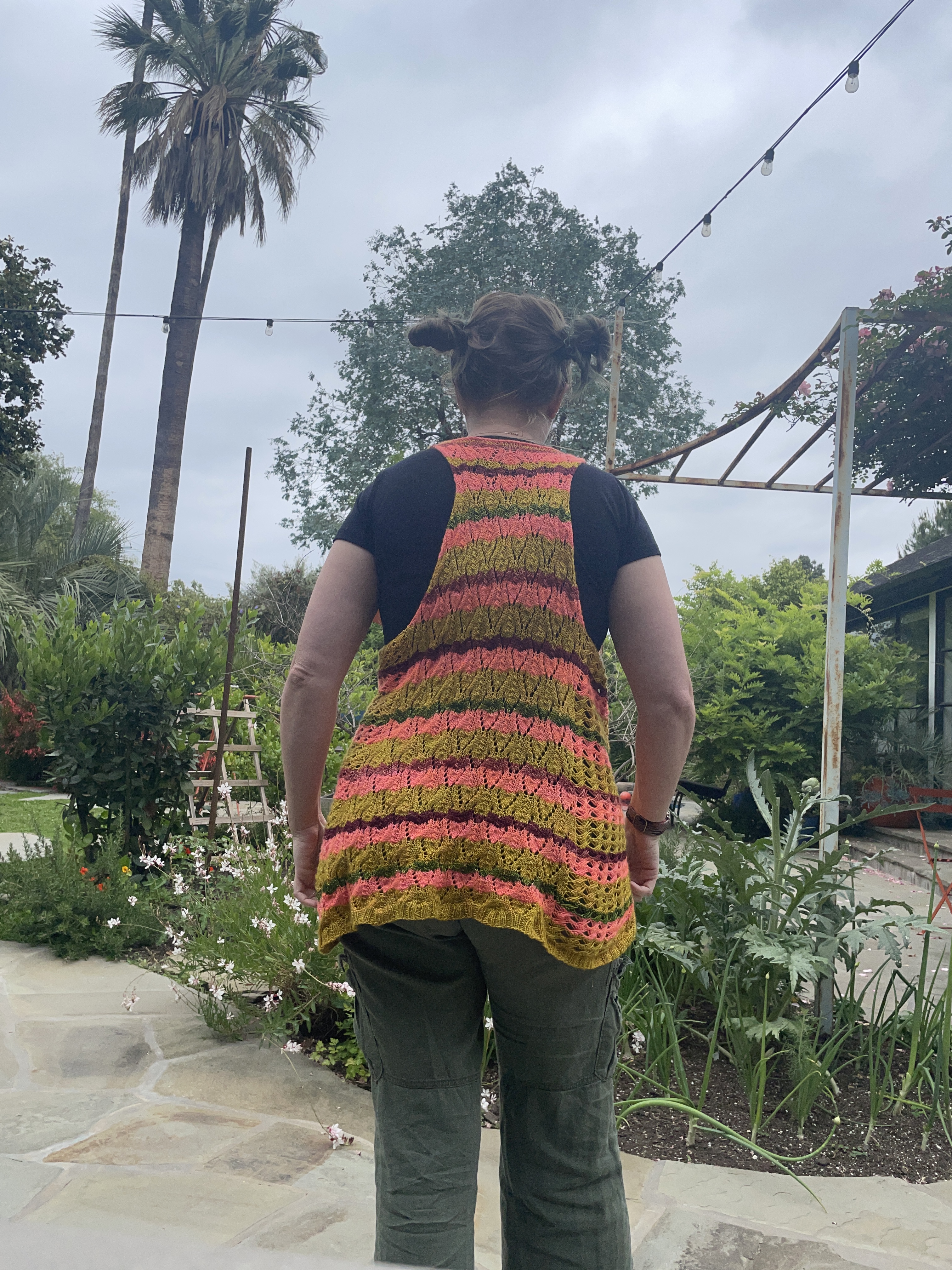 A white woman with her hair up in two high side buns faces away from the camera in a garden wearing a knit striped lace vest in bright yellow, orange, red, and green