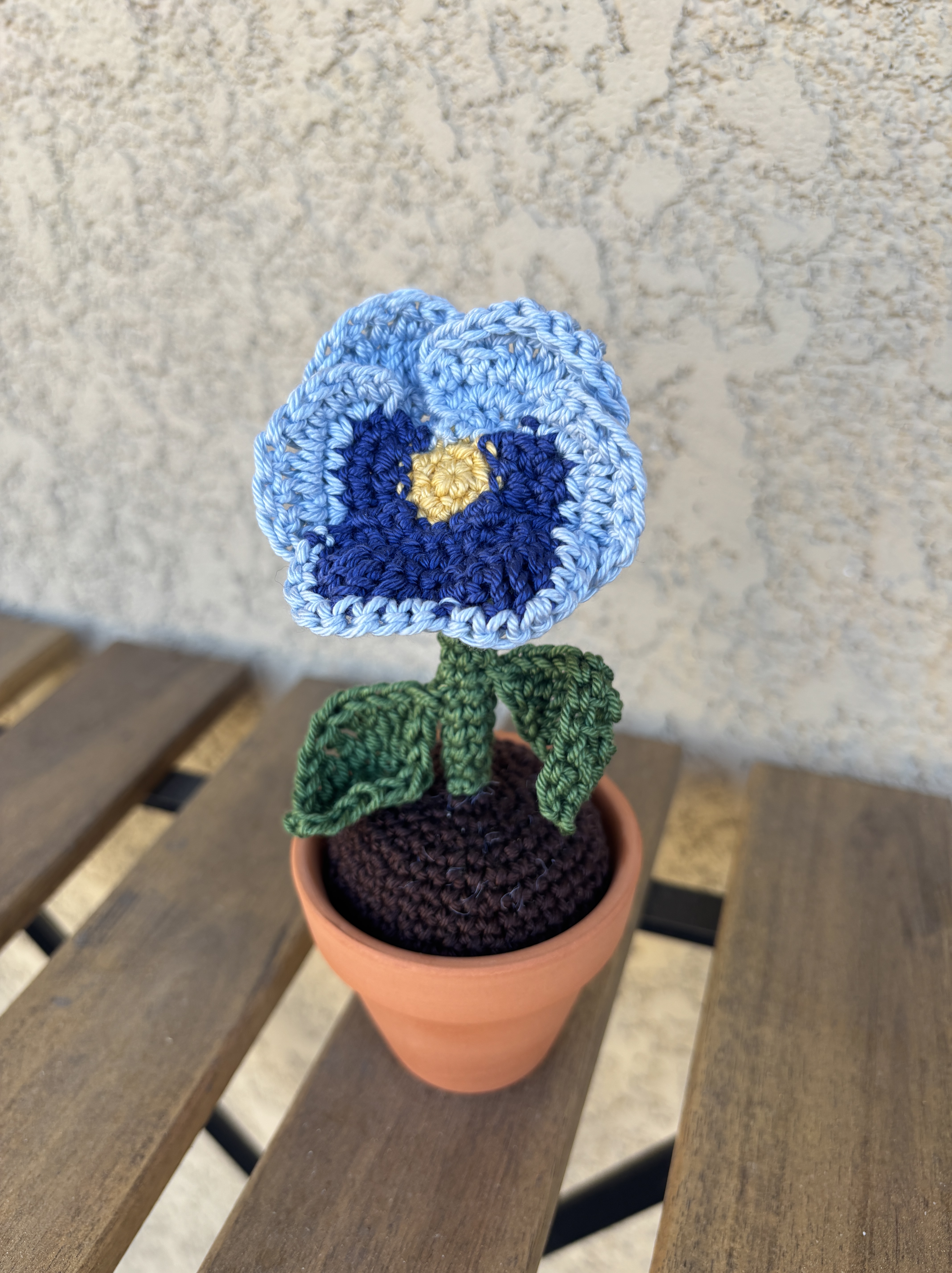 A crocheted pansy in a tiny terra cotta pot with dark brown crocheted dirt, dark green stem and leaves, and light and dark blue petals on an outdoor table
