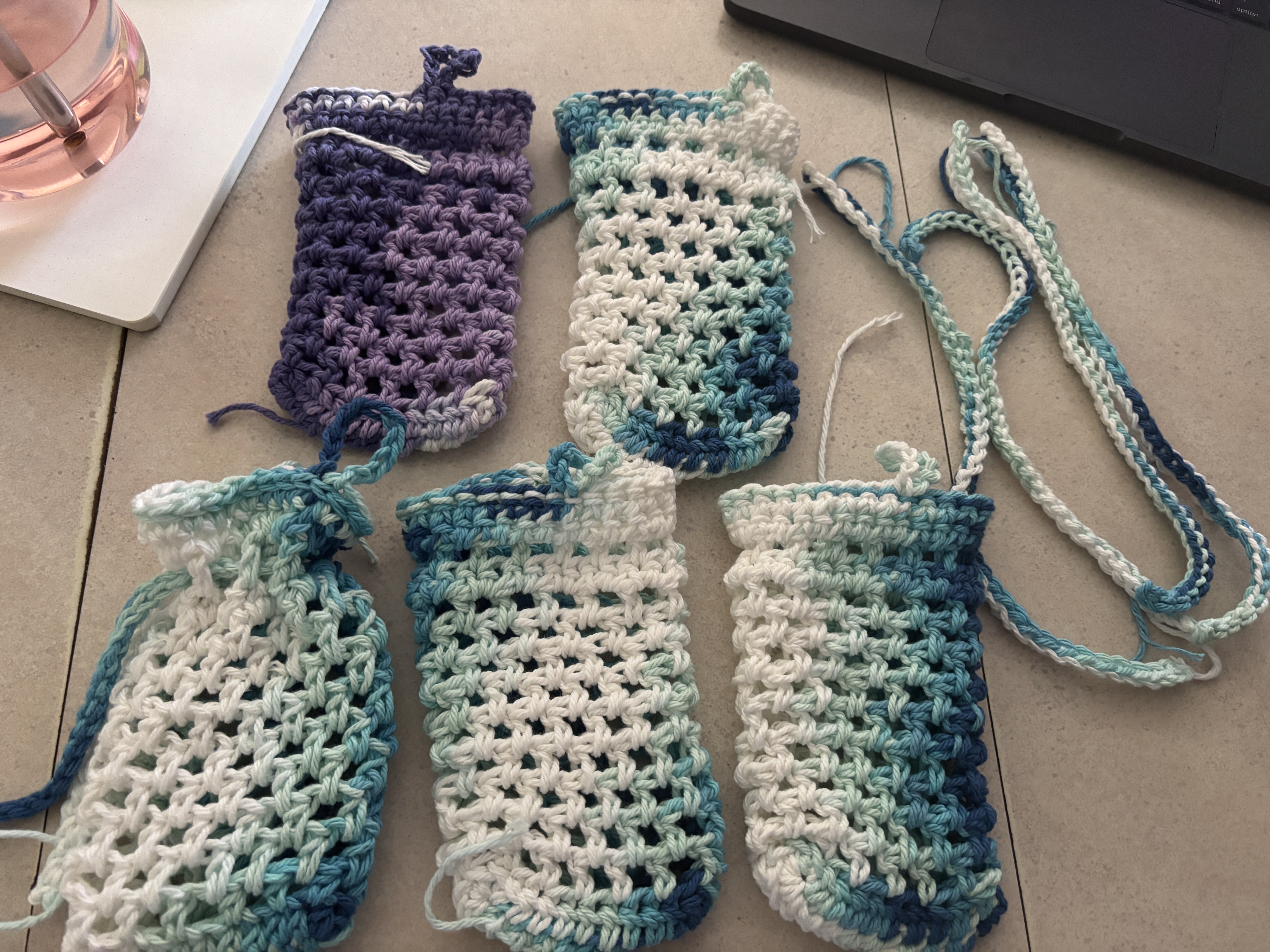 Five crocheted soap sacks with mesh stitch patterns on a pink table. The top left is in shades of purple nad the rest are gradients of blue to white.