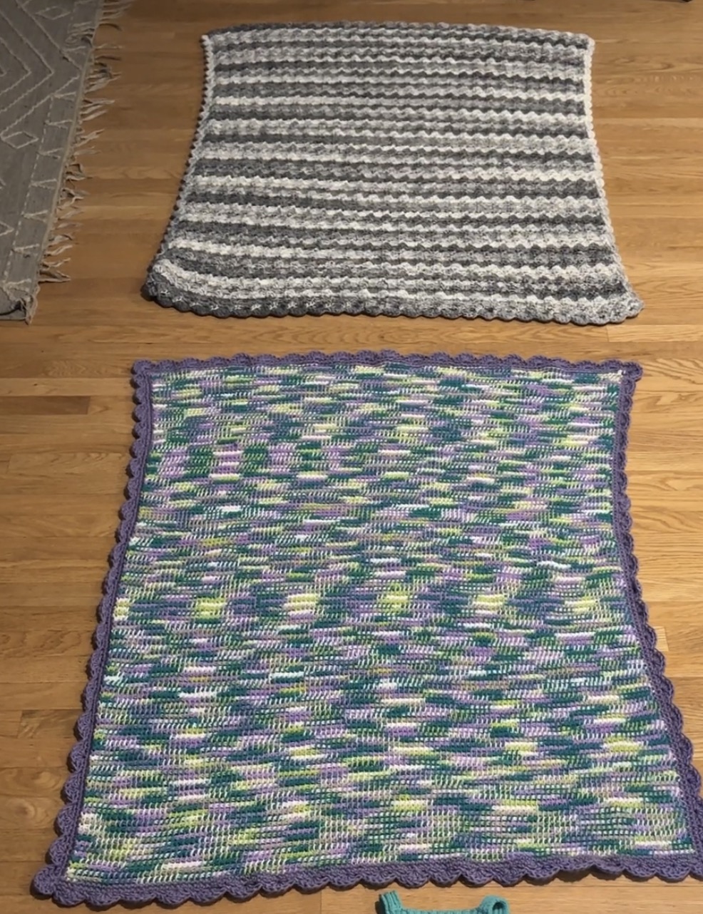 Two crocheted blankets laying on a light wooden floor. The top one is gray and white striped with a diagonally tiling stitch pattern and the bottom is Tunisian crocheted in a purple, green, and white variegated yarn with a solid purple scalloped border